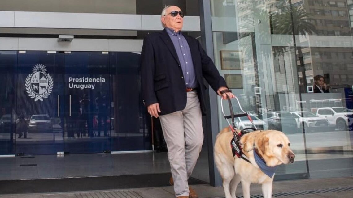 Alberto Calcagno sale del edificio de Presidencia de Uruguay, en Montevideo, acompañado de su perro guía. Lleva gafas de sol y sostiene el arnés del perro mientras ambos avanzan por la entrada del edificio.