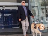 Alberto Calcagno sale del edificio de Presidencia de Uruguay, en Montevideo, acompañado de su perro guía. Lleva gafas de sol y sostiene el arnés del perro mientras ambos avanzan por la entrada del edificio.