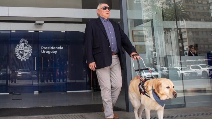 Alberto Calcagno sale del edificio de Presidencia de Uruguay, en Montevideo, acompañado de su perro guía. Lleva gafas de sol y sostiene el arnés del perro mientras ambos avanzan por la entrada del edificio.